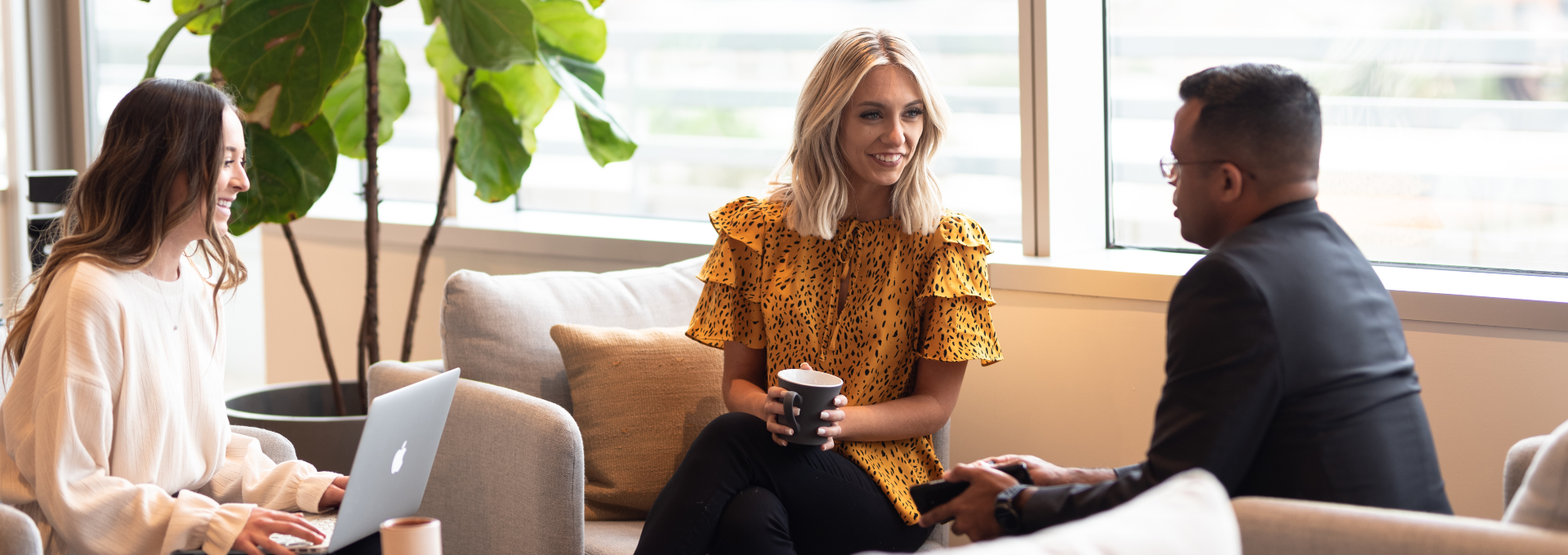 Two women and a man are gathered a small coffee table talking. One woman is working on her laptop while the others are looking at each other.