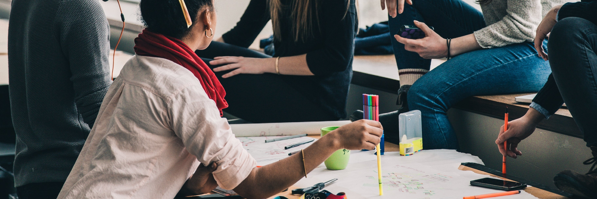 A group of students surround a work table. A large piece of paper covers most of the table. The students are talking to each other.