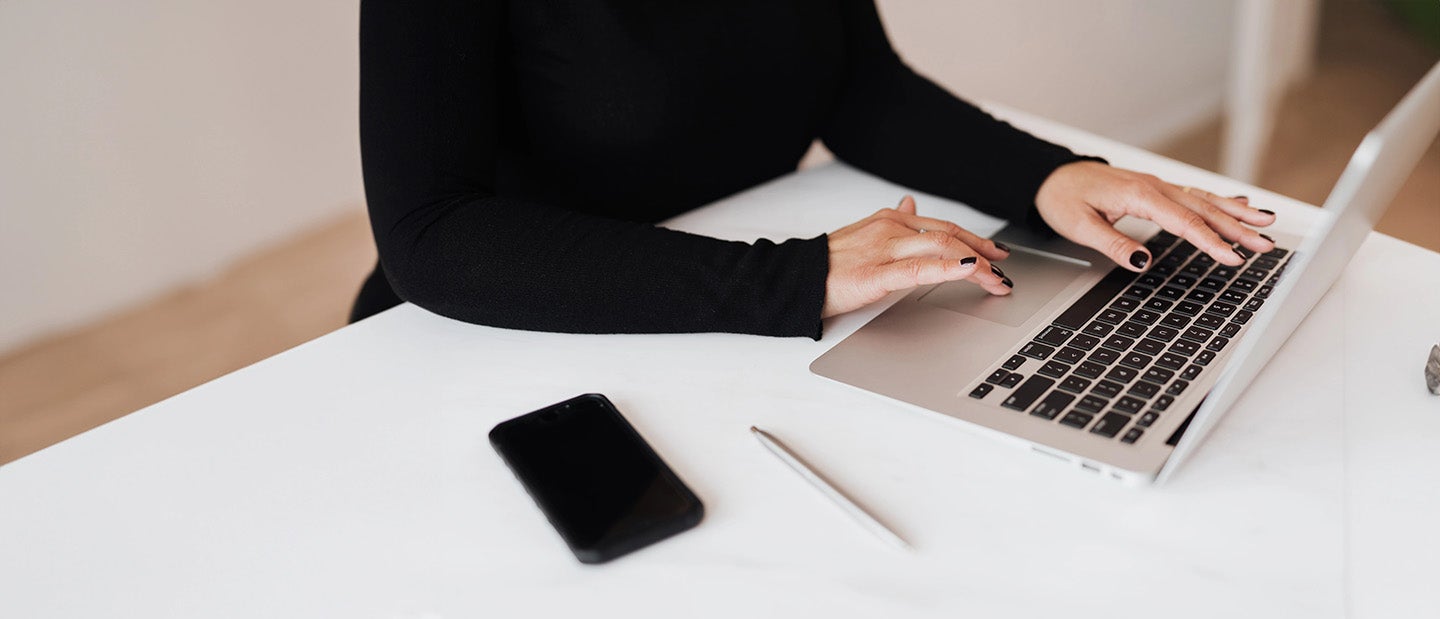 Person typing on a laptop at a white desk with a smartphone and pen.