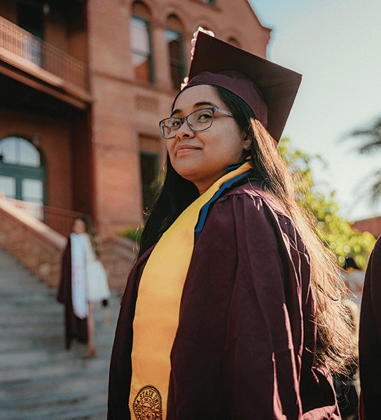 Cindy Rogel Bahena, a graduate in a maroon cap and gown with a yellow stole stands in front of a brick building.